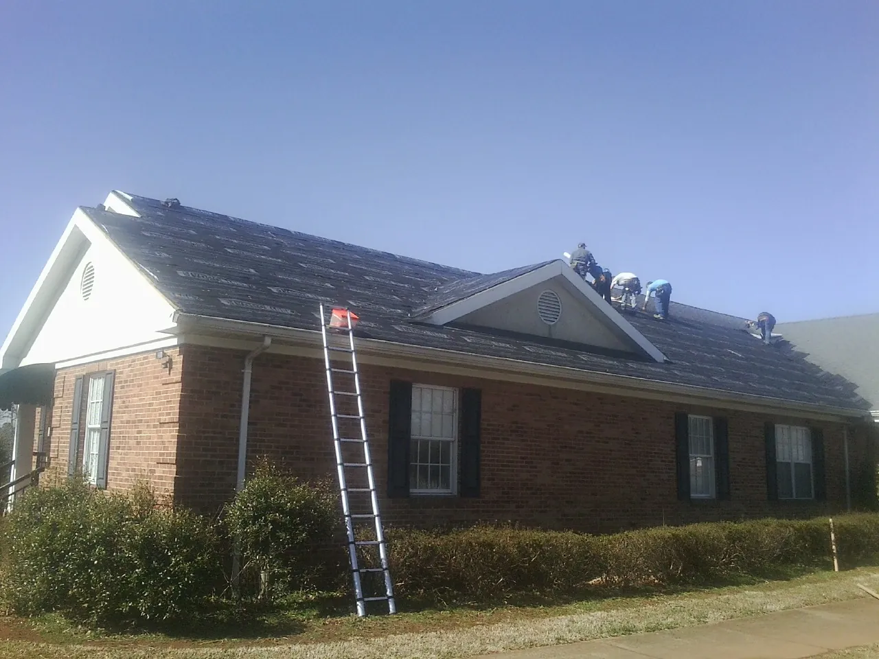 Crew tearing shingles off a brick ranch roof with a ladder set — Emergency Roof Repair work by our crew in Ledgewood