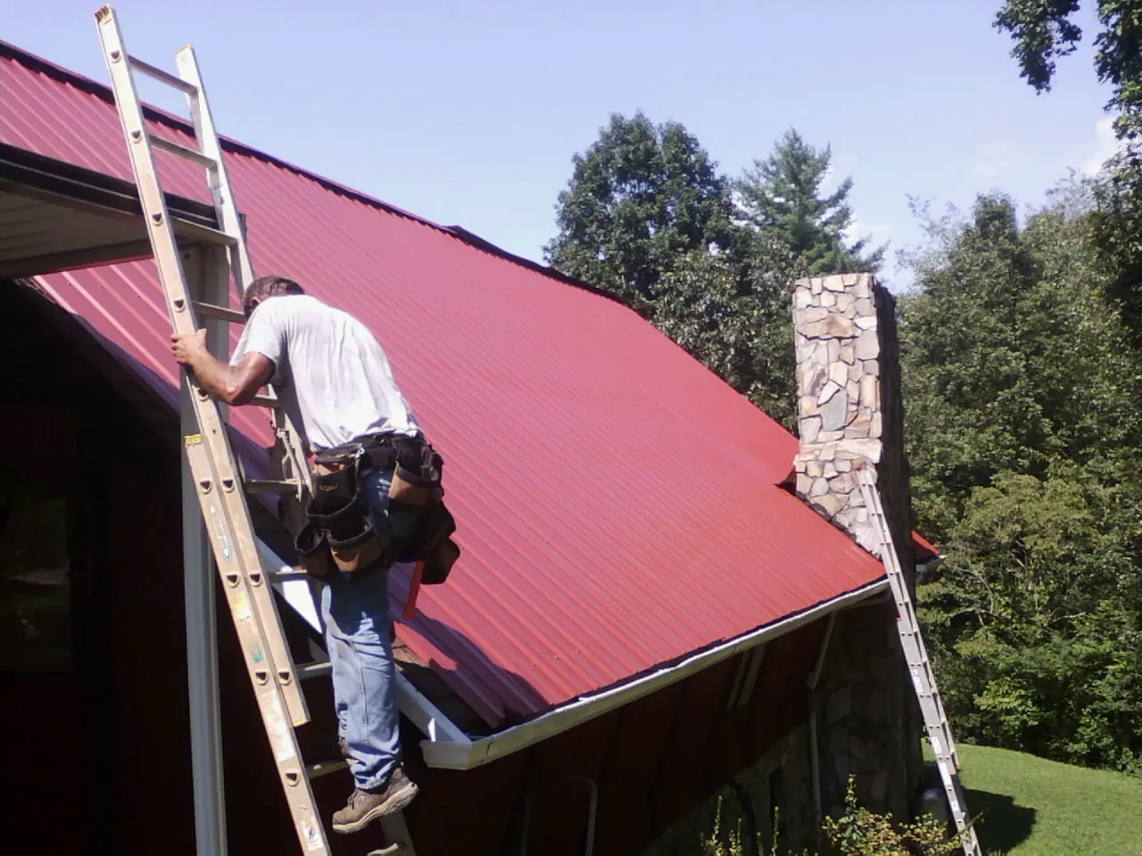 Roofer on a ladder beside a red metal roof and stone chimney — Soffit & Fascia Repair by Roofing Brothers in Ledgewood